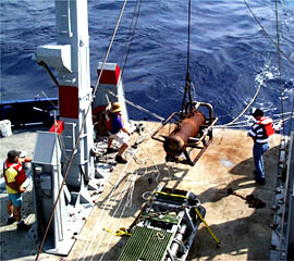 Once the DSL-120 sonar “fish” has been deployed, the clump weight is lowered off the fantail. Ron Comer directs the operation while Randy Dickau (left) and Jeff Keeler (right) man the tether lines on either side of the ship’s A-frame. 