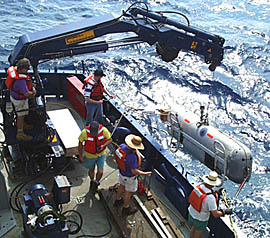 The WHOI team, helped by the Scripps shipboard technicians, lowers the DSL-120 sonar “fish” into the water today to start the survey of our third study area. PJ Bernard is at the crane controls, Jeff Keeler holds the stern tag line on the sonar fish, and Bob “Yogi” Elder keeps the nose of the sonar fish steady. Ron Comer (yellow shirt) lets the Mate on the Bridge know that the fish is over the side, and Randy Dickau (purple shirt) coils up the lines. 