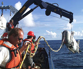 The DSL-120 sonar system is lowered over the side for the last DSL-120 lowering of this expedition. Bob “Yogi” Elder (foreground) and Craig Elder (middle) hold the fiber optic tether line that connects the sonar fish to the clump weight. Randy Dickau hauls in one of the tether lines used to hold the DSL-120 steady during the lowering. PJ Bernard is operating the crane. 