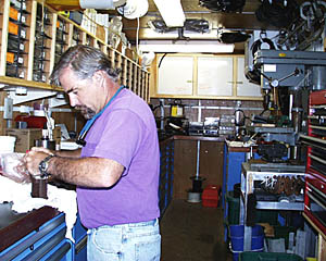 PJ Bernard at work in the WHOI Shop Van. The Van is a container usually used on ships to hold cargo, that has been transformed into a working space. The Van holds many of the spare parts and supplies that the WHOI technicians need to make repairs to their equipment. All the basic tools are stored in the blue cabinets beneath the workbench. The plastic trays by PJ’s head contain small mechanical fasteners, spare parts, electrical connectors, and other items. They are all numbered and cross referenced to lists of parts that are constantly updated. That way, when the cruise is over, the Van can be resupplied easily. 