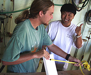 Graduate students Robbie Young and Yong-Jin Won make numbered markers that will be placed at each sampled hydrothermal vent. The white rectangle markers are made of a special material called syntactic foam. The three-foot yellow line attaches the marker to an anchor. Yong-Jin flashes a ‘thumb’s up’ because the line fits through the hole he drilled.  