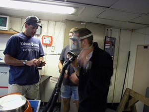 Alvin pilot Bruce Strickrott guides Dive and Discover photographer Ian Yurdin in the finer points of donning an oxygen mask. This would only be necessary if a serious problem arose with Alvin. The challenge is to form a perfect seal between mask and face, because any leak could endanger the lives of all three people in the submersible. 