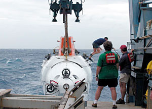 Expedition Leader Pat Hickey, in green shirt, and Alvin Pilot Anthony Tarantino watch as Alvin is raised back on board. Alvin Technician and Pilot-in-training Mark Spear, who acted as a swimmer for this recovery, jumps from the sub to the churning sea. He was then picked up by the Avon nearby.  